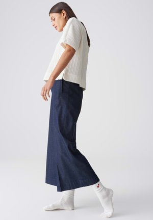 Femme portant un haut en maille blanche ample, un pantalon large bleu marine et des chaussettes blanches avec des accents en forme de cœur rouge, debout avec un pied légèrement levé.