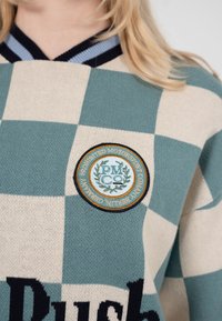 Person iført en grøn og creme ternet sweater med et rundt broderet mærke, hvor der står "Prohibited Motorsport Company Berlin Germany."