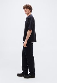 Νεαρός άνδρας με σγουρά μαλλιά στέκεται πλαγίως φοράει χαλαρό μαύρο πόλο μπλουζάκι, μαύρο παντελόνι και μαύρα παπούτσια slip-on σε λευκό φόντο.