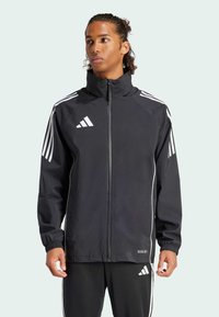 Schwarze Zip-Jacke mit hohem Kragen, weißem Adidas-Logo und drei weißen Streifen an den Ärmeln. Verfügt über elastische Ärmelbündchen und ein leichtes Material.