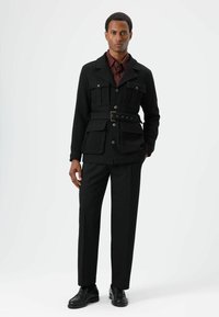 Zwart gecoate jas met meerdere zakken en een ceintuur in de taille, gedragen over een bordeauxrood overhemd en zwarte broek, aangevuld met zwarte schoenen.