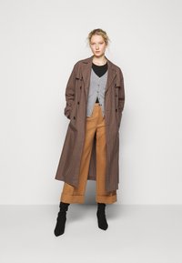 Brun og beige rutete trenchcoat, grå knappet cardigan, svart topp, brune vide bukser og svarte høye hæler.