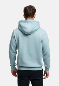 Ljusblå hoodie i mjukt tyg, med stor luva, ribbade muddar och fåll, sedd bakifrån. Mörka byxor syns i nederkant.