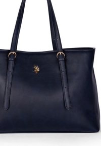Granatowa skórzana torba ze złotymi elementami, z dwiema długimi uchwytami i gładką teksturą. Logo umieszczone jest na froncie torby.