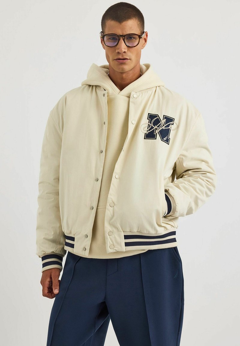 Beżowa kurtka bomberka z granatowymi paskami przy mankietach i dolnym obszyciu, z haftowanym logo na piersi, noszona na jasnym hoodie.