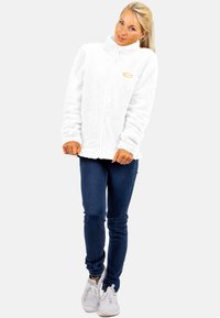 Witte, fluffy fleece zip-jas met een hoge kraag. Heeft een subtiele gele logo op de borst. Gecombineerd met donkerblauwe jeans en witte sneakers.