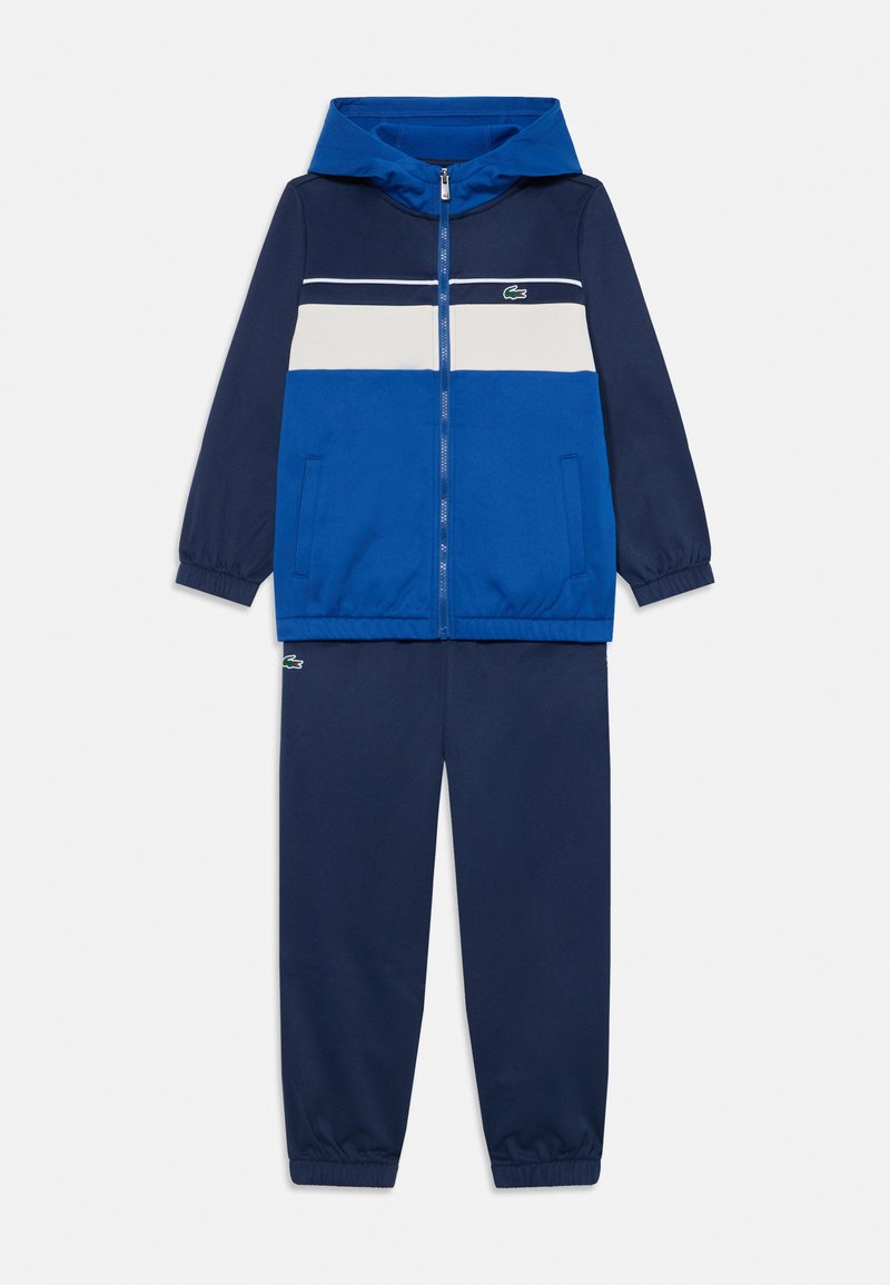 Blå träningsdräkt som har en zip-up jacka med huva, kontrasterande ränder, och matchande byxor med elastiska muddar, gjord av mjukt tyg.