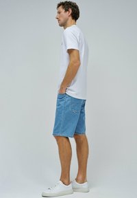 Shorts de mezclilla azul claro con corte recto y bolsillo trasero, combinados con una camiseta blanca lisa y zapatillas blancas, mostrados en vista lateral.