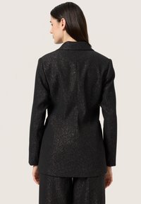 Fekete blézer fémes fényű, struktúrált vállakkal, egy gombos zárással és enyhén hosszabb hátrésszel. Sima anyag, finom textúrával.