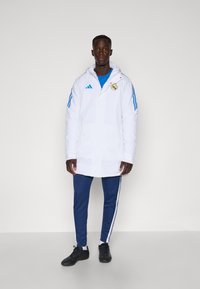 White long jacket with hood and blue accents, featuring the Real Madrid logo. Paired with blue pants and black athletic shoes.