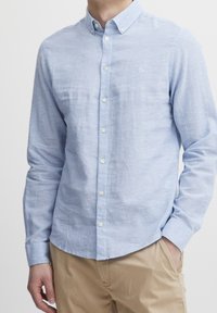 Mies, joka on pukeutunut vaaleansiniseen napapaidan, jossa on logo rinnassa, ja vaaleisiin housuihin. Hänellä on yksi käsi taskussa yksinkertaista valkoista taustaa vasten.