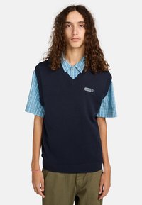 Navy gebreide vest met een V-hals, met ribbelboorden aan de onderkant en armsgaten, gedragen over een blauw geruite overhemd met knopen. Inclusief een logo patch aan de voorkant.