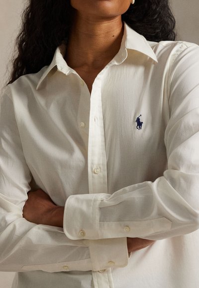 Personne portant une chemise blanche boutonnée, cheveux foncés, bras croisés, montrant le logo bleu du joueur de polo sur la poitrine.