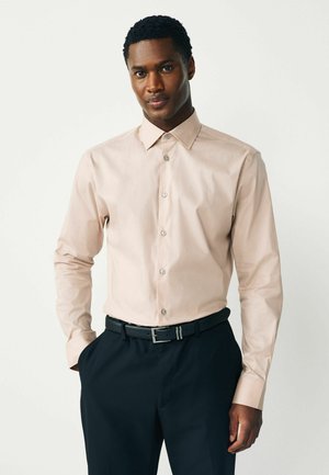 Muž nosiaci béžovú košeľu na gombíky zastrčenú do čiernych elegantných nohavíc s čiernym opaskom, stojaci s jednou rukou v vrecku.