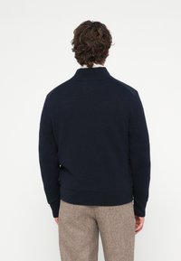 Tumši zila džemperis ar augstu apkakli, ribotām aprocēm un apakšu; rakstainas plecu detaļas un mīksts audums. Bikses gaiši brūnā rūtiņā.