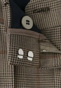 Mankiet brązowo-beżowej kurtki w kratę z dwoma srebrnymi spinkami do mankietów i dużym beżowym guzikiem na czarnej podszewce.