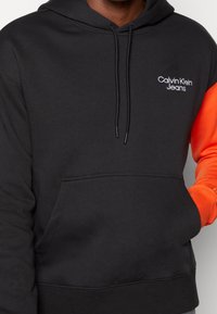Sudadera negra con mangas naranjas, hecha de tejido suave. Cuenta con un bolsillo delantero y el logo de Calvin Klein Jeans en blanco en el pecho.