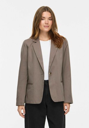 Kvinde med langt brunt hår iført en taupe blazer over en hvid top og løstsiddende sorte bukser, stående mod en ensfarvet lys baggrund.