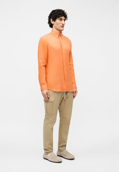 Άνδρας που στέκεται φορώντας πορτοκαλί πουκάμισο με μακριά μανίκια και κουμπιά, μπεζ παντελόνι cargo και μπεζ μοκασίνια σε ένα απλό λευκό φόντο.