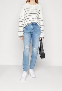 Biały sweter dzianinowy z czarnymi paskami, zestawiony z jasnoniebieskimi jeansami z przetarciami i białymi trampkami. Trzyma czarną torebkę.