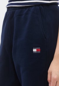 Μοντέρνο παντελόνι φόρμας σε ναυτικό μπλε, κατασκευασμένο από βαμβακερό μείγμα ύφασμα, με κεντημένη πιετά "Tommy Jeans" σε κόκκινο, λευκό και μπλε στη μία πλευρά.