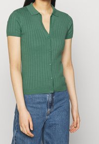 Modell bär en grön ribbad kortärmad cardigan med knappar, i kombination med blå högmidjade jeans, mot en grå bakgrund.