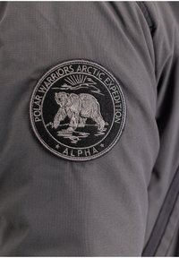 Szürke szövetdzseki, kör alakú fekete folttal, amelyen egy hímzett jegesmedve, nap és a "POLAR WARRIORS ARCTIC EXPEDITION ALPHA" felirat látható.