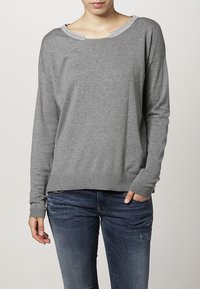 Grauer Pullover mit langen Ärmeln und weitem Ausschnitt, verziert mit einem glitzernden Saum. Aus weichem Material mit gerippten Bündchen und Saum. Getragen mit Jeans.