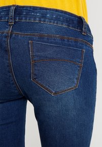 Mörkblå jeans med en klassisk femficksdesign, kontrasterande orange sömmar och en slät textur. Midjan är medelhög.