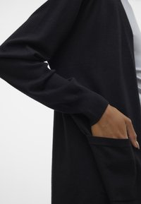 Zwarte lange mouwen cardigan gemaakt van zacht materiaal, met een zak aan de linkerkant. De textuur is glad met geribbelde manchetten.