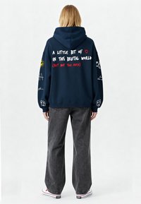 Felpa blu navy con grande testo grafico sul retro. Presenta lettere bianche e rosse, e dettagli neri sulle maniche. Abbinata a pantaloni grigi a gamba larga.