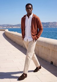 Homme portant un sweat à capuche marron, une chemise blanche, un pantalon beige et des mocassins foncés, marchant le long d'une promenade en bord de mer par une journée ensoleillée.