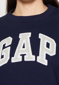 Sudadera azul marino con el logotipo "GAP" en letras bordadas de color blanco y gris claro. Presenta un cuello redondo clásico y una textura suave.