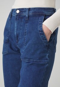 Džínové kalhoty v tmavě modré barvě, s vysokým pasem, předními kapsami a hladkou strukturou. Látka vykazuje jemné variace v tónu.