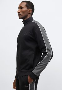 Čierna bunda na zips s šedými detailmi, s vysokým golierom, bočnými vreckami a bielym lemovaním pozdĺž rukávov. Logo na hrudi.