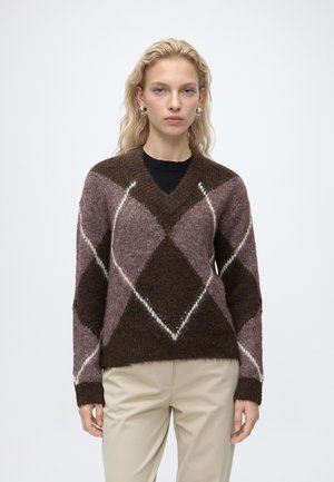 Donna con capelli biondi mossi che indossa orecchini a cerchio d'argento, un maglione a rombi marrone e viola e pantaloni beige, in piedi davanti a uno sfondo semplice.