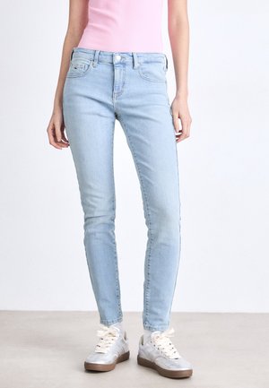 Vrouw die lichtblauwe skinny jeans, witte veter sneakers en een roze mouwloze top draagt tegen een effen achtergrond.