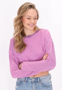 Giovane donna con lunghi capelli biondi mossi, che indossa un crop top rosa a maniche lunghe e jeans blu, sorridente e che guarda in basso con le braccia incrociate.