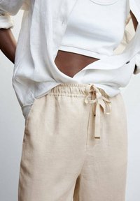 Beige linnen broek met elastische tailleband en decoratieve strik. Gecombineerd met een witte cropped tanktop en een lichte, luchtige overhemd met knopen.