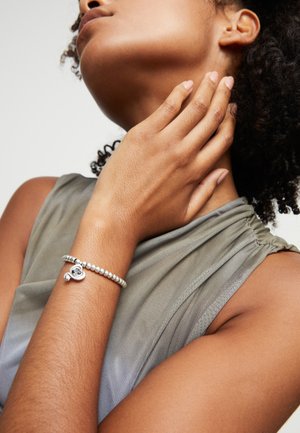 Pulsera de cuentas de plata con un charm en forma de corazón, llevada en una mano descansando sobre una clavícula. El fondo es neutro y suave.