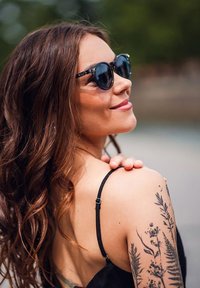 Žena s dlouhými vlnitými vlasy, která nosí sluneční brýle a černý top na ramínka, na jejím horním paži se nachází podrobná botanická tetování.
