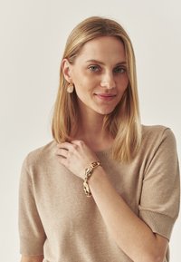 Beige kötött pulóver rövid ujjal. Arany karkötő csavart mintával a csuklón. Arany ovális fülbevalók az arc mellett.