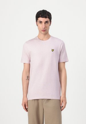 Joven con cabello corto y oscuro que lleva una camiseta rosa claro de manga corta con un pequeño logo de un pájaro amarillo en el pecho y pantalones beige.