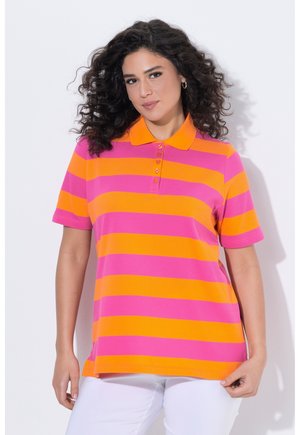 Kvinde med krøllet hår iført en kortærmet polotrøje med brede horisontale lyserøde og orange striber og hvide bukser.