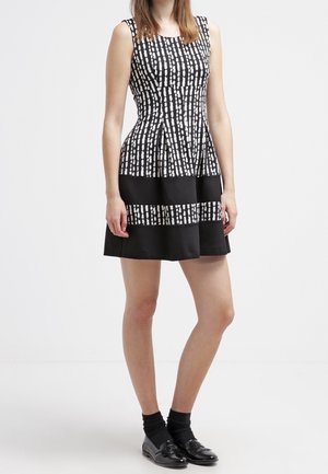 Femme portant une robe noire sans manches avec des motifs carrés blancs et des bandes noires horizontales, assortie à des chaussettes noires et des mocassins.