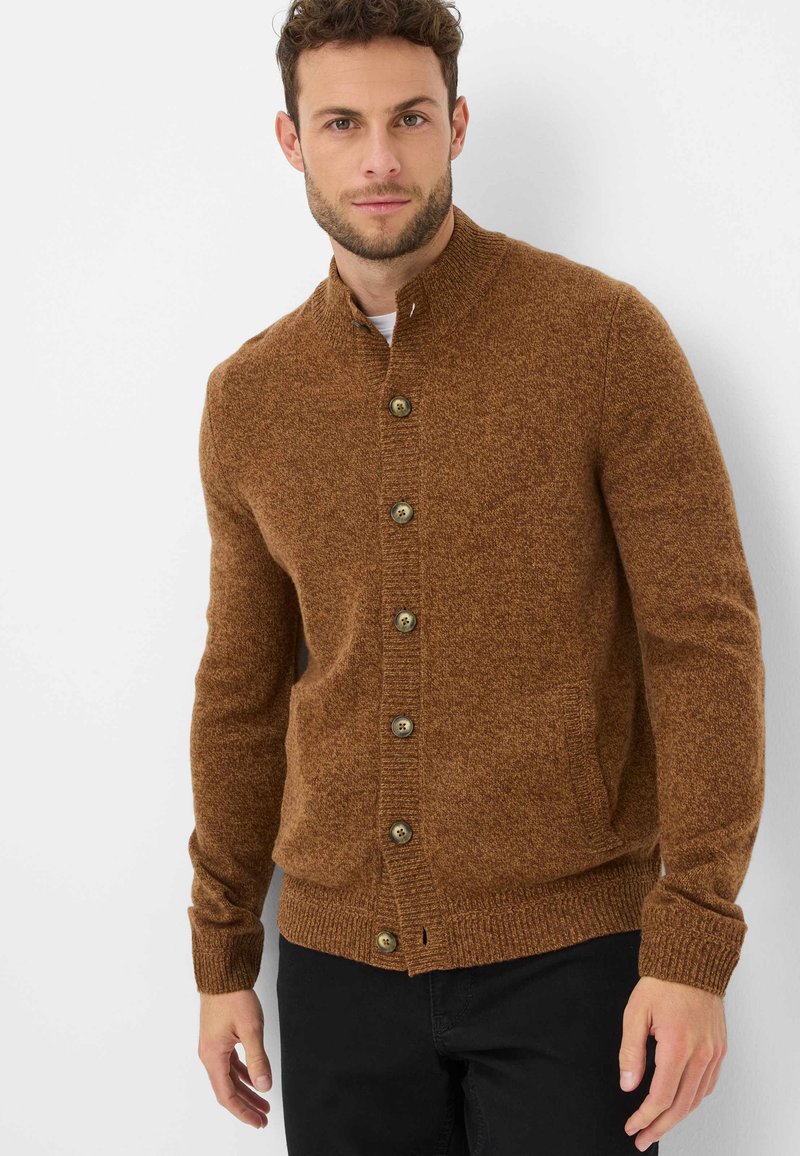Cardigan en tricot marron avec un col haut, un tissu texturé, huit gros boutons et des poignets et un ourlet côtelés, porté sur une chemise blanche.