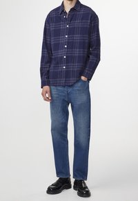 Marineblauwe geruite overhemden met witte knopen, gecombineerd met lichtblauwe rechte jeans en zwarte leren schoenen, staande tegen een witte achtergrond.