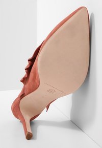 Spitze pinke Wildlederschuh mit High Heels, schräg gegen eine weiße Wand gelehnt, zeigt die glatte Sohle mit Markenname und Größenangaben.