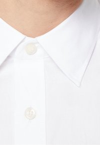 Biała bawełniana koszula z klasycznym, spiczastym kołnierzykiem, centralnym guzikiem i gładką fakturą. Tkanina wygląda na świeżą i starannie wykończoną.