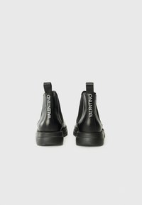 Bottes noires en cuir à la cheville avec des panneaux élastiques sur les côtés, semelle en caoutchouc plate et logo sur la lanière du talon. Texture lisse. Design minimaliste.